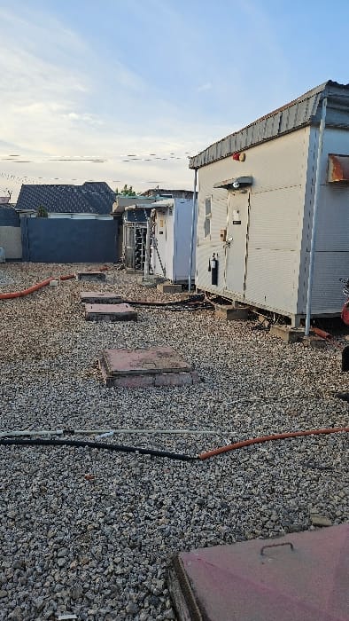 The image displays an outdoor utility or telecommunications site located in Kabala, Nigeria. The ground surface is covered with light grey and brown gravel. Several cables and conduits, including orange, black, and grey varieties, are routed across the gravel. Four reddish-brown, rectangular access covers are visible embedded in the gravel, leading towards a row of structures. These structures are modular, pre-fabricated-style shelters, light-colored (off-white/light grey), with corrugated metal roofs, elevated on concrete bases. The closest shelter on the right features a white access door, a small window or vent, and an attached red device, possibly a fire extinguisher. Further along the row, other shelters and exposed equipment, potentially cooling units or electronic cabinets, are visible. In the background, a dark-colored building with a sloped roof is partially observed. The sky is light blue with sparse white clouds. No individuals are present in the scene.