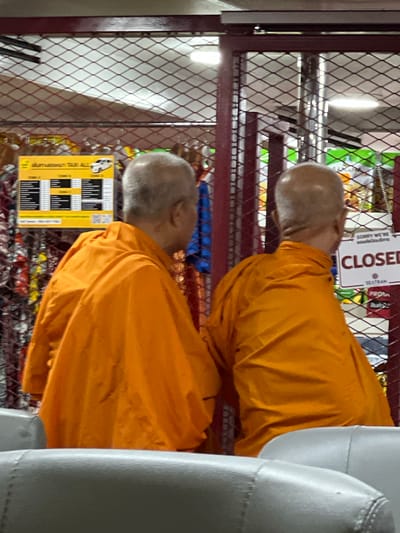 This media file captures two individuals, likely monks given their orange robes and shaved heads. They are positioned within a setting that appears to be a store or a waiting area, as indicated by the wire shelving filled with various items and a sign that reads "CLOSED". The overall lighting suggests it might be daytime, though the interior setting limits definitive conclusions about natural light.
One monk is seen from the back, with his head tilted down. The other monk is visible from the side and slightly from the front. The clothing of both individuals is a distinct saffron orange, a common color for Buddhist monks' robes. The background shows shelves with products, some appearing to be food items or packaged goods, and a blue fabric is visible beneath the shelving. A prominent sign in English and Thai states "CLOSED" and "SORRY WE'RE". Another smaller sign below it includes Thai text and a table-like structure with headings like "TAXI ALL" and "ZONE".
The scene appears to be static, with no active event taking place beyond the presence of the individuals and the displayed information. The overall mood is neutral, with no strong indicators of emotion. The presence of the "CLOSED" sign suggests that whatever business or establishment this is, it is not currently operational. The location is hinted at by the Thai language on the signs, suggesting a setting within Thailand. The presence of seating, possibly from a waiting area, is also subtly visible on the right side of the frame.