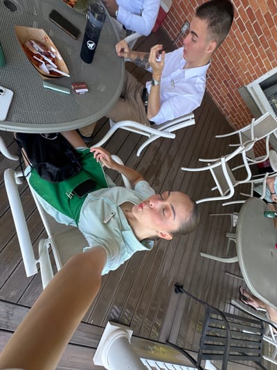 The image captures an outdoor gathering on a wooden deck. In the foreground, a person, possibly a woman, is lying down and taking a selfie, puckering her lips. She is wearing a light blue and white striped shirt and green shorts. A black drawstring bag rests on her lap.
In the background, a man in a white collared shirt and khaki pants is seated at a round table. He appears to be gesturing with his hand. The table has a brown paper container with utensils, a black water bottle, and a small rectangular object on it.
Several white outdoor chairs are scattered around the deck. A brick wall is visible in the upper right portion of the image. The wooden deck appears to be wet in some areas. The overall scene suggests a casual outdoor social event. The provided crops offer closer views of the table items and the individuals. The location is identified as Harbor Springs, United States of America.