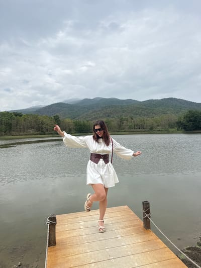 A young woman poses on a wooden dock overlooking a lake. The dock is constructed of light-colored planks and has rope ties to wooden posts at the edges. The lake reflects the overcast sky. Behind the lake is a forested mountain. The woman is wearing a white dress with a brown belt, sandals, and sunglasses. She is standing with her arms outstretched. The location is Ban Huai Sai, Thailand.