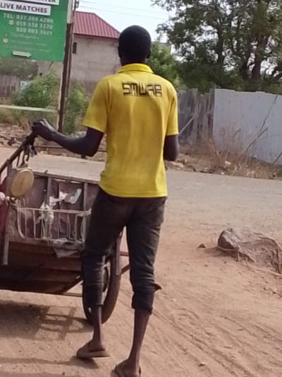 A full-length, back-view shot captures an individual standing next to a two-wheeled cart on a dirt path. The person, likely male, is dressed in a bright yellow short-sleeved polo shirt and dark trousers that appear cropped above the ankles. He is holding the handlebars of the rustic, wooden-framed cart, which contains various items or possibly discarded materials. His bare feet are visible, clad in simple sandals.

The setting suggests a rural or semi-urban environment, consistent with Dungu, Ghana. The ground is unpaved, made of dirt and sand, with some scattered rocks. In the background, there is a mix of natural elements and structures. Dense green foliage and trees are visible, along with a distant building featuring a prominent red roof. A wooden fence runs along the right side of the path.

A large, green informational sign is partially visible on the left side of the frame. Clearly legible text on the sign includes "LIVE MATCHES" at the top. Below this, several telephone numbers are listed: "TEL: 037 209 4290", "059 938 3370", and "020 003 36f1". An arrow points to the right.

The back of the individual's yellow shirt displays block-letter text, which appears to read "SMUAB". The image is taken in bright daylight, indicating either morning or early afternoon, with clear weather. No specific emotions are discernible from this angle. The scene depicts a common sight of daily life, possibly involving a vendor or transporter, in a Ghanaian community.