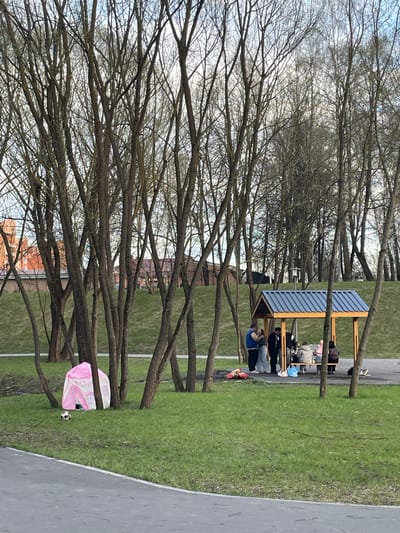 The image shows a park in Fabriki 1 maya, Russia. In the background, a group of people are sitting on benches under a covered shelter. The foreground is dominated by leafless trees, their branches creating a dense network across the sky. Near the bottom of the frame, on the grass, is a pink and white play tent with a soccer ball nearby. A paved path runs along the right edge of the image. The overall impression is of an early spring day with people enjoying the park despite the bare trees.