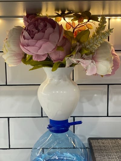 A bouquet of artificial flowers, including large pink and white peonies, is arranged in a white ceramic vase. The vase is precariously balanced on the neck of a large, clear blue plastic bottle, secured by a blue plastic cap or band. The background consists of a white tiled wall with black grout lines, and a shelf or surface above the flowers is illuminated by warm LED lights. The scene appears to be indoors, possibly in a kitchen or bathroom, given the tiled wall. The location is Dolgoprudny, Russia.