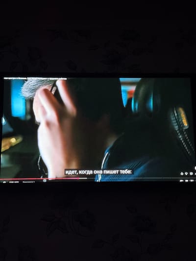 This image captures a television screen displaying video content within a dimly lit room in Ciorescu, Moldova. The screen shows a medium-close shot of a person from behind, with their right hand raised near their head, seemingly touching their grey or light-colored hair, which is styled upwards. The background of the video content is blurred, featuring subtle blue and yellow ambient lighting. Along the bottom of the screen, white Russian subtitles are present, reading: "идет, когда она пишет тебе:" (translating to "goes when she writes to you:"). Below these subtitles, standard video player controls, including a red progress bar and various icons, are visible, indicating the video is hosted on a platform resembling YouTube. Faint Russian text is also discernible at the top left of the screen, referencing "центры в Молдове. Kоторые попадают в трудовое рабство" (translating to "centers in Moldova. Who fall into forced labor"). The environment outside the television screen is dark, revealing a wall covered with dark wallpaper featuring a subtle floral or leafy pattern.