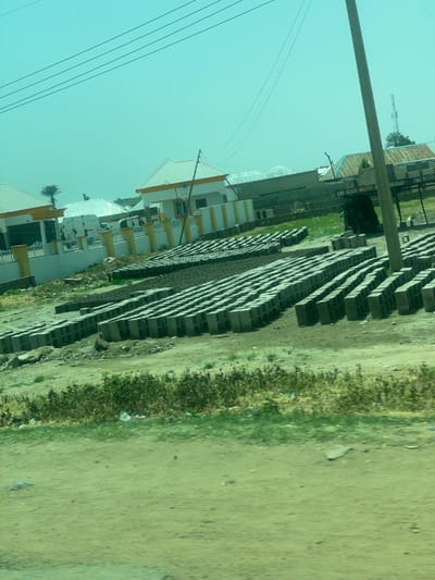 The image shows an outdoor scene, likely in Bauchi, Nigeria, with a clear, pale blue sky dominating the upper portion. In the foreground, a thick, gray horizontal beam, possibly part of a structure or utility pole, stretches across the image. To the left of the beam are several parallel black electrical wires.
Below the beam, a row of multi-story buildings with white and yellow exteriors are visible. These buildings appear to be residential or commercial, with balconies and windows. Some have white, sloped roofs. A palm tree can be seen peeking out from behind one of the buildings.
To the right of the buildings is a large area filled with neatly stacked gray concrete blocks, arranged in long rows. This suggests a construction site or a block manufacturing facility. Beyond the blocks, there is a patch of green grass and vegetation, and then a dirt or gravel road. The scene appears to be lit by daylight, with no strong shadows to indicate a specific time of day. The overall impression is one of everyday life in a built-up area with industrial or construction activity.
Text is visible on one of the buildings in the background, on a dark vertical surface. It reads "KMK BLOCKS INDUSTRY N". Another smaller sign above it seems to read "BEROAD".