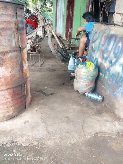 A cluttered outdoor garage scene reveals a person sitting with their back turned, wearing a blue shirt and dark shorts. Beside them are bottles and a large plastic container filled with snack wrappers. An old motorcycle in disrepair is positioned nearby. A rusted metal barrel stands to the left, and a graffitied wall forms the backdrop on the right, completing the disorganized workspace.