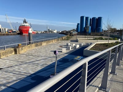 The image depicts a waterfront walkway in Gorleston-on-Sea, United Kingdom, featuring a paved area with a blue artificial surface and interactive playground elements, including square concrete blocks, a purple standing microphone device, and a blue spinning object. A modern metal railing runs along the right side of the walkway. Beyond the walkway, there is a concrete dock with a red offshore supply vessel docked along with large white tanks and a crane. In the background, there are a series of tall blue storage tanks. The sky is clear and blue.