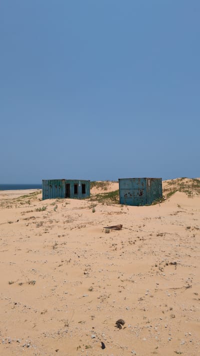 The image shows a sandy beach with two stacked blue shipping containers in Phuoc Dinh, Vietnam. The containers are rusty and appear abandoned. The sky is clear blue. The beach is sparsely vegetated.