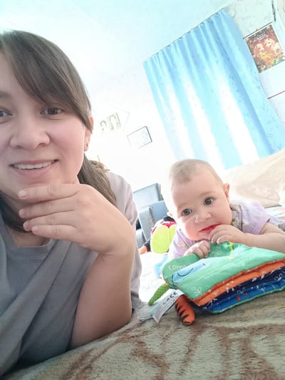 A woman and a baby are lying on the floor in a room. The woman is in the foreground, looking at the camera and smiling. She has dark hair and is wearing a gray shirt. Her left hand is resting on her chin. The baby is to her right, also lying on the floor. The baby has light hair and is wearing a purple shirt. The baby is looking at the camera and has a red object in its mouth. In front of the baby is a stack of colorful fabric books. The room has light blue curtains in the background and what appears to be a calendar on the wall. The floor is covered with a patterned rug. The location is identified as Каменное, Russia.