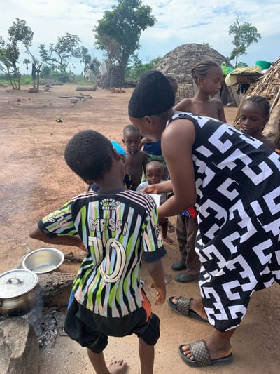 In the city of Nandu, Nigeria, a group of children and an adult woman gather in a dusty, outdoor setting. The woman, wearing a black and white patterned dress, is bent over, interacting with a young boy in a striped jersey with "MESSI 10" on the back. Several other children, boys and girls, stand around them, some looking at the interaction, others appearing more disengaged. In the background, traditional thatched-roof huts and trees dot the landscape under a cloudy sky. Cooking pots are visible on rocks, with smoke rising from one, suggesting a meal is being prepared. The environment appears rural, with dirt ground and minimal infrastructure.