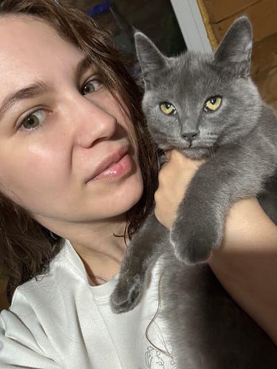 A selfie shows a person with light skin, light brown hair, and hazel eyes holding a gray cat. The person is wearing a white shirt, and a silver chain necklace is visible. The cat has green eyes. The background appears to be an interior space with wooden paneling. No information is available to confirm the location of Nizhnyaya Gar', Russia.