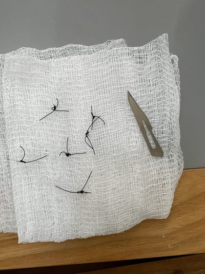 A metal scalpel blade rests on a white gauze pad, which is also scattered with short lengths of black surgical suture. The pad sits partially on a grey surface and partially on a light brown wood surface. The sutures are tied in knots at their midpoints, with loose ends extending outwards. The gauze's woven texture is visible.
