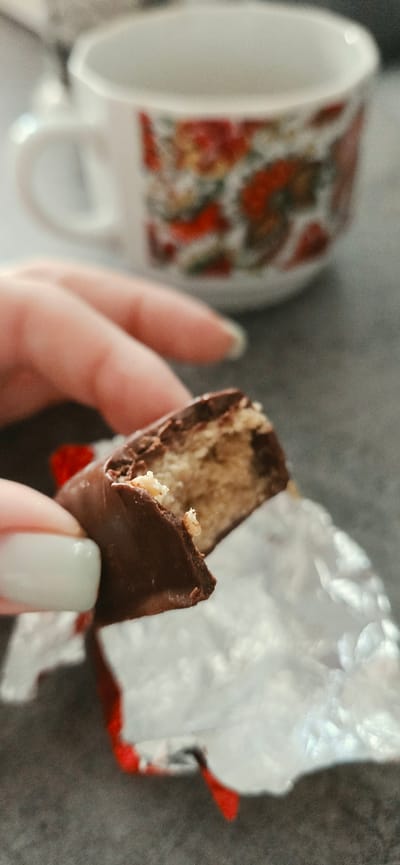 A hand with light-colored nail polish is holding a partially eaten chocolate-covered candy bar, which is partially unwrapped from its foil packaging. In the blurred background, a white mug with a floral pattern is visible, suggesting a domestic or café setting. The scene is set on a textured gray surface. There is no indication of the specific location within Lipetsk, Russia, or any other city.