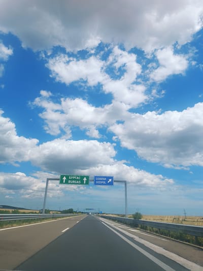 The image shows a view from a car driving on a highway in Bulgarovo, Bulgaria. The highway stretches ahead into the distance, with a guardrail on the right and a grassy median on the left. The sky is a vibrant blue, filled with fluffy white clouds. Two road signs are visible: one green sign with white text and arrows indicating "БУРГАС" and "BURGAS" pointing straight ahead, and a blue sign with white text and an arrow indicating "СОЗОПОЛ" and "SOZOPOL" pointing to the right. The surrounding landscape appears to be rural, with fields of dry grass and scattered trees, and power lines visible in the distance. The sun appears to be high in the sky, suggesting it is daytime, and the weather seems pleasant with clear skies and clouds. There are no people or other vehicles immediately visible in the foreground, and the scene evokes a sense of travel and forward movement.