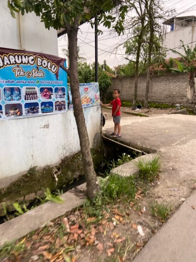 The image shows a young boy in a red shirt and blue shorts standing on a sidewalk in Margaasih, Indonesia. The boy is looking to the left, with his left hand on a wall. The wall is painted white and has a sign advertising a bakery called "Warung Bolu Teh Adah". The sign depicts various kinds of cakes and pastries. The boy is standing next to a large tree with a thick trunk. There is a small, open drainage ditch between the sidewalk and the wall. The ditch is filled with water and small plants. The ground around the ditch is covered in leaves and debris. There is a white building in the background, with other houses visible beyond it. The area is generally quite calm and serene, with few people or vehicles present. The image was taken in Margaasih, Indonesia at the coordinates -6.953 latitude and 107.551 longitude.