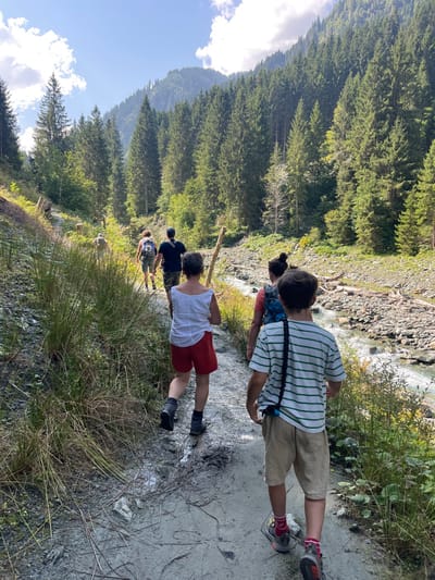 The image depicts six individuals hiking along a dirt path in a forested mountain valley near Reitlehen, Austria. The scene is illuminated by bright daylight under a partially cloudy blue sky. A clear, rocky river flows to the right of the path, with its banks strewn with logs and stones. Dense conifer forests cover the steep mountain slopes on both sides of the valley.

In the foreground, a young male wearing a white and green striped shirt, khaki shorts, and sneakers walks away from the camera; a black elastic strap with a blue object is attached to his side. Behind him, an adult female wears a white sleeveless top, red shorts, and dark boots. Further back in the line, another adult female wears a red top and a blue backpack, followed by an adult male in a dark shirt and camouflage shorts. Two additional individuals are partially visible further along the path. The path itself is uneven, with sections of damp earth and scattered pine needles and small debris. Cut tree stumps are visible on the rising slope to the left of the path.