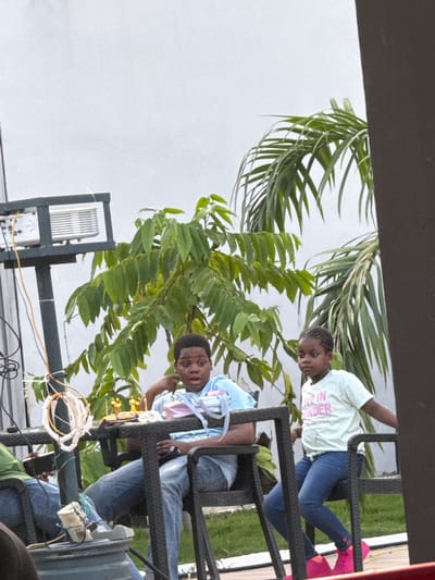 In Rubuchi, Nigeria, two young people are seated outdoors on a dark-colored metal furniture set, possibly a patio or balcony. The person on the left, a young girl with braided hair, wears a light-colored t-shirt with pink text that reads "KEEP IN ORDER" and blue jeans. She is looking downwards, her expression neutral. To her right, a young boy, wearing a light blue t-shirt and blue jeans, is looking at the camera with a slight smile. His hands are resting on the furniture.

The scene is framed by lush green foliage, likely palm leaves and other plants, suggesting a tropical or subtropical environment. The background appears to be a light-colored wall or sky, indicating it is daytime. The overall atmosphere seems relaxed and casual. A piece of equipment, possibly an air conditioning unit or a server rack, is visible in the lower left portion of the image, with some wires connected. The lighting suggests it is either late afternoon or early morning, with soft light.