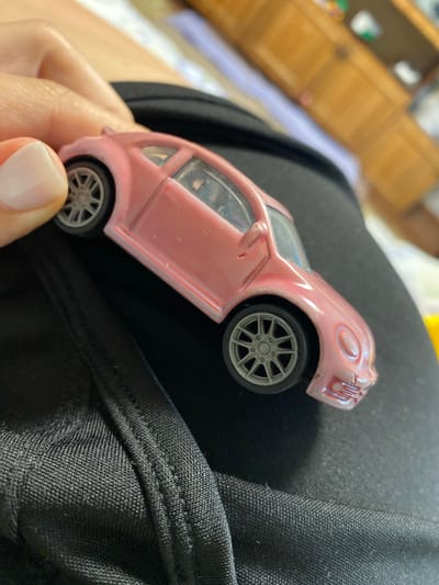 A person's hand with light-colored fingernails is holding a small, pink toy car, likely a Volkswagen Beetle, over a black, textured fabric, possibly clothing. The toy car is facing left and appears to be slightly worn, with some scuff marks visible on its surface. The background is blurred, showing hints of furniture and possibly other objects. There is no indication of the location in Michura, Russia, within the image.