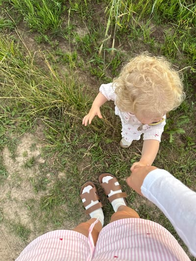 This is a first-person perspective, eye-level shot, taken outdoors on a dirt path with scattered grass. The image features a toddler with curly blonde hair wearing a white dress with a cherry print, white socks, and tan sandals. The toddler is holding the hand of an unseen person. In the foreground, the legs and feet of the unseen person are visible, wearing pink and white striped shorts and brown sandals with white socks. The scene appears to be in a rural or park-like setting. The location is not specified in the image or its metadata.