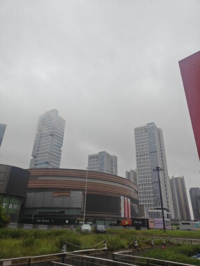 This media file shows a street view of a modern urban environment in Shunde District, China. The scene is dominated by several tall, contemporary buildings with glass facades, indicative of a commercial or business district. One distinctive skyscraper has a unique, stepped design, partially obscured by a light fog or haze, suggesting overcast weather. In the foreground, a large, curved building with a brown, textured exterior, possibly a shopping mall or entertainment complex, is visible. This building features large display screens and banners, one of which displays Chinese text and the word "TARI".

Below the large building, a row of food trucks and temporary structures are parked. One truck is labeled "Pull-Tab" and another "Meet Fresh", suggesting a food or beverage vendor area. Chinese text on banners and signage indicates commercial activity, with phrases like "怡翠地产—广佛品牌房企" (Yicuiprod-Guangfo brand real estate enterprise) and "怡翠晋盛" (Yicuijin sheng). A green barrier with more Chinese text is present in front of the buildings.

To the left of the main structure, a sign with "应急出口" (Emergency Exit) is visible. A patch of green grass and vegetation lines the foreground, with metal railings and a concrete structure suggesting a pedestrian walkway or a drainage system. The overall atmosphere is muted, likely due to the cloudy or foggy weather, which softens the sharp lines of the architecture. No people are prominently visible, giving the impression of a quiet moment in a bustling urban setting. The time of day appears to be daytime, given the ambient light.