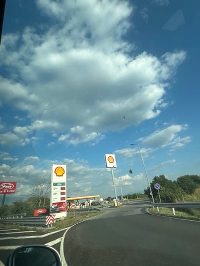 The media file is a photograph taken from a vehicle on a sunny day, likely in the afternoon, with scattered white clouds against a bright blue sky. The perspective is from inside the car, looking outwards through the windshield. The scene is dominated by the sky and clouds, with a roadside view on the right.
Visible elements include a Shell gas station with prominent signage featuring the Shell logo and prices for different types of fuel (e.g., FuelSave V5, FuelSave Diesel, AutoGas) alongside the word "café". There are also signs for "Happy Bar & Grill". Road markings and a guardrail are present, suggesting a highway or main road. A red and white directional sign is also visible. Trees line the background, indicating a rural or semi-rural setting.
The image is oriented vertically, making the sky appear to be on the left and the road on the right. There are some reflections on the windshield, such as a rectangular shape in the upper left corner. The overall mood is bright and casual, suggesting a travel or roadside stop. No people are visible in the frame. The location context is Radinovo, Bulgaria.