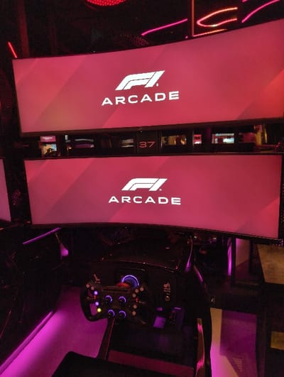 A dimly lit image shows two Formula 1 Arcade booths flanking a race car simulator. The booths, with a reddish-pink hue, display the Formula 1 logo and the word "ARCADE" vertically. One booth has the number 37 on its side. The simulator features a detailed steering wheel with multiple buttons and dials. The scene is illuminated with purple ambient lighting.
