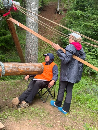 Two individuals are engaged in an outdoor activity in a wooded area. The person in the foreground is seated on a folding chair, wearing an orange safety vest, black pants, and hiking boots. Their head is tilted down, and they appear to be holding something small in their hand. The second person, standing behind and to the left of the first, is wearing a grey raincoat over a red hoodie, black pants, and blue Crocs. They are leaning forward, with one hand resting on a wooden beam, and seem to be looking down.
The scene is set in a forest with lush green foliage and trees. Several ropes and a wide, orange strap are visible, suggesting some kind of setup, possibly for an adventure activity like slacklining or a rope course. A large wooden log is positioned prominently in the foreground, with rope tied around it. Another fallen tree trunk is visible in the background. The ground is covered with dirt and grass. The overall impression is that of a daytime activity, possibly during overcast weather given the muted lighting. No visible text is present in the image. The location is identified as Sergino, Russia.