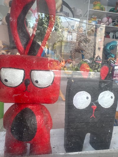 The image shows a display in a shop window, possibly in Givatayim, Israel. The foreground features handmade, cartoonish figures. One is black with two large white eyes and a red snake tongue. The other two are red, also with large white eyes and snake tongues. The red figures are connected, resembling a caterpillar. The backdrop is a distorted reflection of a street scene, including a giraffe statue, buildings, and other indistinct figures and objects. The reflection indicates an urban environment. There are also other figures on shelves inside the shop.