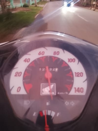 The image shows a close-up of a motorcycle's dashboard as it is being ridden. The speedometer is the central focus, with markings for 0, 20, 40, 60, 80, 100, 120, and 140 km/h. The needle is currently pointing to approximately 32 km/h. Below the speedometer, the "HONDA" logo is visible. There are also indicator lights for the turn signals, showing arrows pointing left and right, and a symbol that may represent the headlight. The background is blurred due to motion, suggesting the motorcycle is in transit. Hints of a road with a white line and some greenery and buildings on the side are visible, indicating an outdoor, possibly rural or suburban, setting. The lighting suggests it could be daytime or early evening. The location context provided is Pante Beurene, Indonesia.