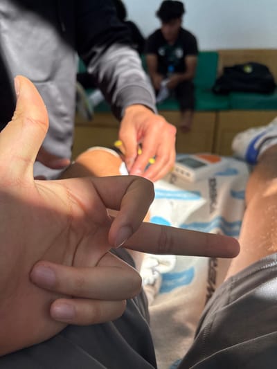 The image shows a close-up of a hand giving the middle finger. A second person is partially visible, seemingly attending to another individual's leg, possibly wrapping or bandaging it. In the background, a third person is sitting down, near a backpack. The overall setting appears to be an indoor space, perhaps a locker room or training area.
