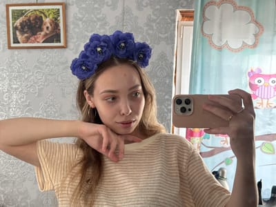 A woman is taking a selfie in a room. She is wearing a light yellow and brown striped shirt, gold hoop earrings, and a blue floral headband with small skull accents. She is holding a light pink smartphone in her right hand, capturing her reflection in a mirror. Her left hand is resting under her chin.
The room has wallpaper with a light grey and white floral pattern. A framed picture of two puppies hangs on the wall. To the right of the woman, a curtain with a cartoon-like owl and cloud design is visible. Other objects are partially visible in the mirror, including a white cylindrical object with what appears to be a lamp or light fixture. The specific location in Кемуль, Russia is not discernible from the image.