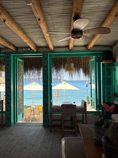 The media file depicts an interior view of a beachfront establishment, likely a cafe or restaurant, in Bodrum, Turkey. The scene is viewed from an angled perspective, with a ceiling fan made of wood and metal prominently featured in the foreground. The ceiling is constructed from interwoven reeds or straw, supported by thick wooden beams.
The establishment opens out to a sunny beach and the clear blue sea. Through the open doorways, beach umbrellas and empty chairs are visible on the sand. In the distance, a person is seen paddleboarding on the water. The decor features vibrant teal-painted wooden frames around the openings, adorned with colorful, decorative tiles.
Inside, wooden tables and chairs with patterned upholstery are arranged. A potted cactus and a plant are placed on a table. The overall atmosphere suggests a relaxed, seaside setting, likely during daytime due to the bright sunlight and clear skies. The image conveys a sense of tranquility and vacation. There is no visible text in the media file.