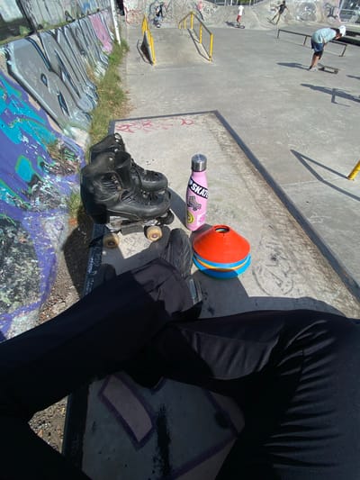 The image is taken at a skatepark in Monte Grande, Argentina. In the foreground, a person wearing black pants and black shoes is sitting on the edge of the graffiti-covered skatepark bowl. Next to the person is a pair of black roller skates resting on a skateboard, a pink water bottle with stickers, and a colorful orange and blue cone. In the background, there are other people skating in the park, with yellow rails and various graffiti designs on the concrete surfaces. The overall environment suggests an outdoor, recreational space.