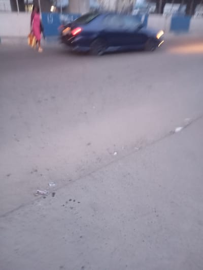 A low-angle, motion-blurred image captures a street scene in Port Harcourt, Nigeria. In the upper left, a person in a pink dress is walking, carrying bags. To the right of the person, a dark-colored car is in motion, with its brake lights illuminated, suggesting it is stopping or slowing down. The car appears to be a sedan. The environment consists of a paved road with some debris scattered on its surface. In the background, there are blurred structures that indicate an urban setting. The lighting suggests it is either dusk or dawn, or a generally overcast day.