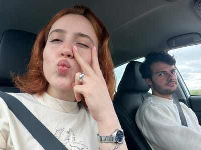 The image is an upside-down selfie taken from inside a car. A red-haired woman with fair skin is in the foreground, making a kissy face and holding two fingers up to her eye. She wears a silver watch and ring, and is wearing a cream-colored t-shirt. Behind her and to the left, a man with dark hair and a beard looks at the camera. Both occupants are wearing seatbelts. The sky is visible through the car window. It is unknown if this was taken in Tedotsminda, Georgia.