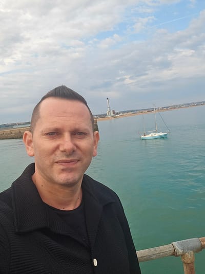 A man in a black jacket and t-shirt stands on a pier, taking a selfie. He has short, dark hair styled into a quiff and a light complexion. Behind him, a pale blue sailboat is anchored in the turquoise water. In the distance, a coastal town with buildings and a tall smokestack can be seen along the shoreline. The sky is overcast with scattered clouds. The scene is likely the coastal area of Adur, United Kingdom, given the context.