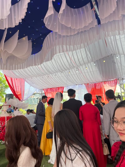 This image captures an outdoor celebration, likely a wedding, taking place under an elaborately decorated canopy in Tay Son Commune, Vietnam. The scene is filled with guests, many dressed in formal attire.
The canopy ceiling is a prominent feature, adorned with cascading white ruffled fabric. Sections of dark blue fabric embedded with numerous small fairy lights create a star-like effect, while panels of red and white striped fabric add to the festive decor. Beneath this, a diverse group of people gathers. In the immediate foreground, a woman with long dark hair and glasses smiles faintly towards the camera. She wears a grey top, and next to her, another person in a traditional red Vietnamese Ao Dai is visible from the back, holding a beige handbag.
Further back, a bride in a white wedding gown and veil is seen from behind, accompanied by a guest in a bright yellow dress. Several men in suits, including a presumed groom in a black suit and a guest in a grey shirt holding a camera, are also present. The event appears to be held on a green surface, possibly turf or artificial grass. Natural daylight indicates it's daytime, while the presence of a fan suggests warm weather. Some green foliage is visible outside the canopy, and artificial red and blue floral arrangements adorn a stand or table. The overall atmosphere is one of celebration and joyous gathering. No discernible text is visible in the image.