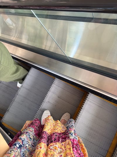 A high-angle shot shows two people riding an escalator. One person is wearing light green pants and a white shoe that is visible in the reflection on the glass wall. The other person is wearing a colorful dress and woven sandals. The setting appears to be an escalator within a building in the United States of America city of Coral Gables.