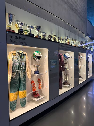The image displays a section of a museum exhibit, likely within the Mercedes-Benz Museum in Stuttgart, Germany, given the prominent display of racing memorabilia. The exhibit features several glass display cases showcasing racing suits, helmets, gloves, shoes, and numerous trophies. In the foreground, a racing suit with green, teal, and yellow coloring hangs on display, accompanied by orange racing gloves and red racing shoes. To the right, a white and green racing helmet with the "BP" logo and the name "STEVE PARRISH" is visible, along with a trophy. Another display case to the right shows a maroon racing suit, a helmet, gloves, and shoes, with trophies displayed behind. Further to the right, another display case contains a racing helmet and other racing equipment. Above the display cases, a row of trophies of various sizes and designs are arranged on shelves. The background shows a concrete wall with engraved text, possibly names of champions or significant figures in racing history. The lighting in the display cases is focused and bright, illuminating the artifacts within. The overall environment appears to be a modern museum with polished floors and sleek display structures.