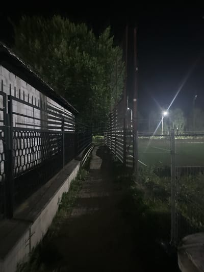 The image depicts a nighttime view of an outdoor area, likely a sports field or park, in Strășeni, Moldova. On the left, a white brick wall runs along a dark metal fence. Beyond the fence and wall, large, dark green trees are visible against the night sky. A narrow, dirt path runs between the wall and fence on the left and another fence on the right. The fence on the right encloses a green, artificial turf field, possibly a soccer or football pitch, illuminated by bright stadium lights. Two tall, wooden posts stand near the fence on the right. The ground around the path and field is covered in dark soil and grass. The scene is dark, with artificial lighting providing the primary illumination. There are no people visible in the image.