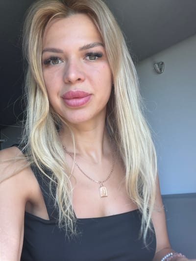 A close-up selfie shows a fair-skinned woman with blonde hair, styled in loose waves. She has prominent false eyelashes, light brown eyes, and full lips with pink lipstick. She is wearing a black tank top and a rose gold necklace with a pendant. The background is softly blurred, with a grey wall to the right, which has a small metal fixture attached to it. The lighting suggests an indoor setting with natural light coming from the left. The image does not contain any information that would allow for its location to be identified as Mykulychyn, Ukraine.
