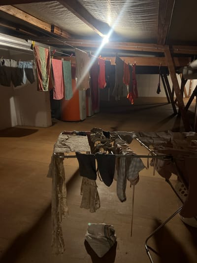 This image captures a dimly lit indoor scene, likely a basement or attic, with wooden beams and textured walls visible. The primary focus is on laundry that has been hung to dry. There are two distinct drying setups. On the left, a clothesline is strung between wooden supports, adorned with various garments, including towels of different colors (red, green, striped) and t-shirts. Some are held by clothespins, both pink and yellow. A bright light source, possibly a lamp, illuminates this area, creating a strong glare.

To the right, a metal drying rack stands on the floor, also holding a collection of clothes. These appear to be lighter fabrics, some with a checkered pattern, and a dark fabric item. Some of the clothes are draped over the rack, while others are suspended. The floor is a warm, brown tone. The overall impression is of a practical, domestic activity taking place in a somewhat utilitarian space. There are no people visible in the image. The time of day and weather are not discernible from the image, but the indoor setting suggests it could be anytime. The location context provided is Mühldorf a. Inn, Germany, which adds a geographical detail to the scene.