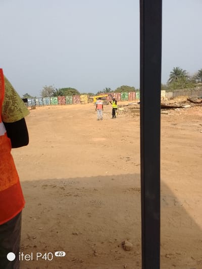 This image depicts an outdoor industrial or storage yard in Hoche, Nigeria. The sky is clear and light-colored. The ground consists of unpaved, dry, light brown soil with scattered small rocks.

In the immediate foreground on the left, a person is partially visible, wearing an orange high-visibility vest over a light green patterned shirt and dark sleeves. A dark vertical metal pole or beam partially obstructs the right side of the foreground.

In the midground, two individuals are present. One person, positioned to the left, is seen from behind, wearing a red-orange high-visibility vest over a light shirt and light-colored pants. To their right, another person wears a bright yellow high-visibility vest over dark clothing and has their right arm raised in a gesturing motion, directed towards the first individual.

In the background, multiple rows of stacked shipping containers of various colors (blue, green, red, brown) are arranged horizontally across the scene. A large yellow piece of heavy machinery, possibly a telehandler or forklift, is positioned to the left of the two midground individuals and in front of the containers. To the far right, a pile of debris, including what appears to be wood and metal, is visible. Beyond the containers and debris, several green trees, including palm trees, are present under the distant horizon.