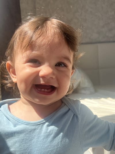 A close-up, canted, full-face shot depicts a young child with light brown hair and brown eyes, smiling broadly with mouth open, revealing teeth. The child is wearing a light blue, long-sleeved shirt. The background is indistinct but suggests an interior space with natural light. Location not specified.