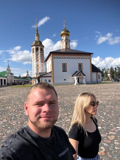 This is a selfie taken by a man in the foreground, with a woman to his right and behind him. They are standing in a cobblestone plaza in front of a white building with a tall, ornate bell tower. The building has a gold dome and is decorated with colorful patterns. Behind the main building are more structures, some with green roofs, and a few trees. The sky is blue with scattered white clouds. People can be seen walking in the background. This appears to be the Suzdal Kremlin in Suzdal, Russia, a historic fortified complex that houses several churches and other buildings.