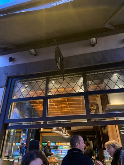 The image captures a storefront scene at what appears to be evening or night, looking into a brightly lit establishment from the darker exterior. The building's exterior wall is light-colored, featuring a square LED floodlight and a hanging circular sign with a green motif. Part of a white sign with glowing blue text is visible on the far left.
Through large multi-paned windows, a diverse interior is visible. The window glass has a distinctive diamond-patterned grid. Inside, several people are present. A man in a dark top is seen in profile, looking towards a blonde woman whose head is partially visible. Further back, another person in a jacket and hat is seated at a counter. In the foreground, the top of a head with dark hair is visible in the bottom right.
The interior decor includes a wooden staircase with vertical slats, a counter area with several silver-colored dispenser taps, and shelving units displaying colorful items, possibly bottles or packaging, illuminated from behind.
Visible text on a sign inside the window includes "Glaces" (French for "ice creams"), "froi" (likely "froid," French for "cold"), and "5.00," indicating a price. These details suggest the establishment is an ice cream parlor or a cafe/dessert shop. The overall impression is of a casual commercial venue busy with customers.