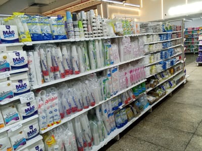 The image shows a supermarket aisle in El Valle del Espíritu Santo, Venezuela, with shelves stocked with various disposable cups and paper products. The items are primarily packaged in clear plastic, with branding visible in colors like blue, white, and red. Specific products include "Más Suave" paper napkins, "Cariocas Servilletas Grandes," and a variety of cups labeled with brands like "Dihamana," "Seli Seli," and "Vaso Soufflé." Some shelves also contain other household items and cleaning supplies. The flooring is tiled with a speckled pattern. There are no people visible in the image. The lighting appears to be artificial, typical of an indoor retail environment.