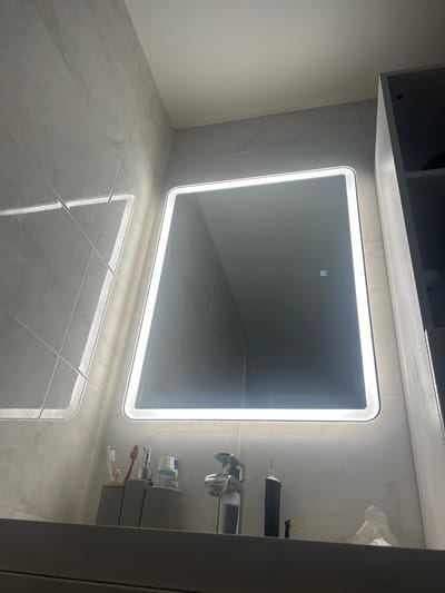 The image shows a modern bathroom with a large, illuminated mirror. The mirror is rectangular with rounded corners and a bright white light strip around its perimeter. In the reflection of the mirror, there is a small square icon with a circle inside. Below the mirror, on the countertop, are several items, including a silver faucet, a grey holder with toothbrushes, a pump dispenser, and a tube of toothpaste. To the right of the faucet, there is a black electric toothbrush with an extra brush head and a crumpled piece of white paper. The walls are tiled with a marble-like pattern. A dark gray cabinet is partially visible on the right side of the image. There are no people present in the scene. The location is identified as Нижние Муллы, Russia.
