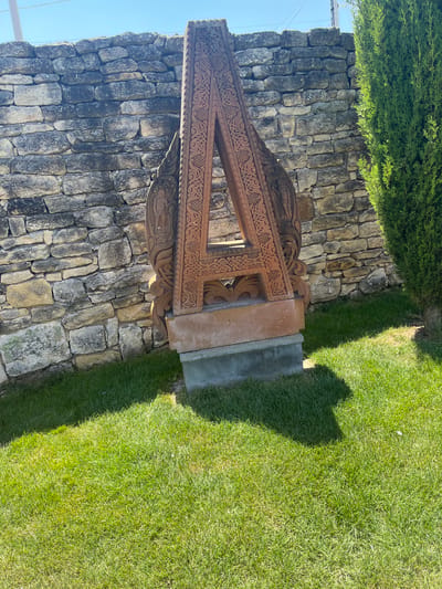 A carved stone khachkar, a traditional Armenian cross-stone, stands outdoors in front of a rustic stone wall. The khachkar is reddish-brown and intricately carved with geometric patterns and depictions of figures, resembling an open book or a triangular structure. It is mounted on a grey base. To the right of the khachkar, a tall, dense green evergreen tree is visible. The ground in front of the khachkar and stone wall is covered in bright green grass, with a distinct shadow cast by the khachkar onto the lawn. The sky above is clear and blue. This khachkar is located in Pliska, Bulgaria.