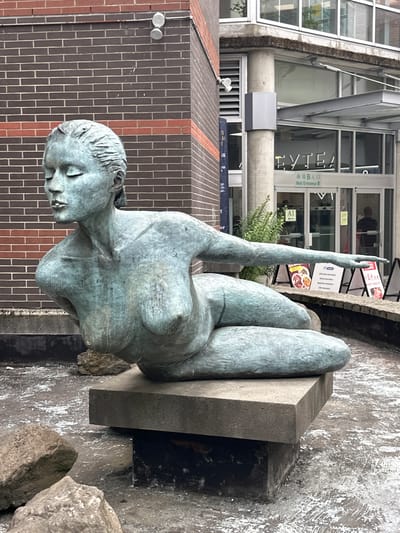 This media file features a bronze sculpture of a reclining woman, positioned on a concrete slab, with her head tilted downwards and eyes closed. Her left arm is raised with the hand pointing upwards. The sculpture has a greenish patina, suggesting it is aged or has been exposed to the elements. The background shows a modern building with large glass windows and a brick wall. Signs are visible in the background, one of which reads "Mall Entrance B" in English and Chinese characters, and another that appears to be an advertisement with food images. The lighting suggests it is daytime, and the overall atmosphere is somewhat tranquil, perhaps reflecting the pose of the sculpture. The setting appears to be an urban environment, possibly a public space or shopping center entrance, given the mall signage.